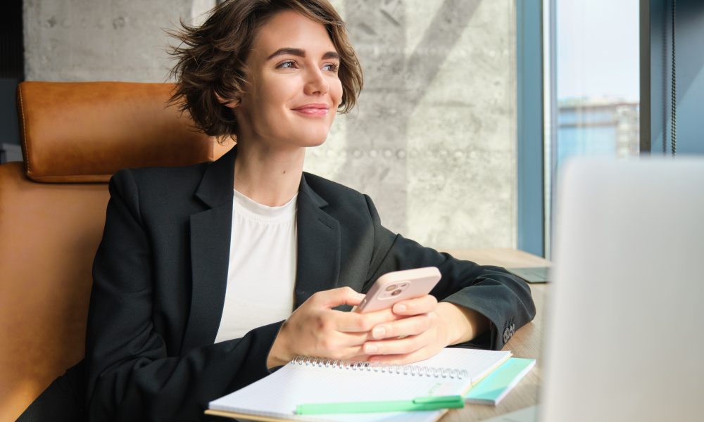 Mulher sorrindo utilizando o celular em um ambiente corporativo